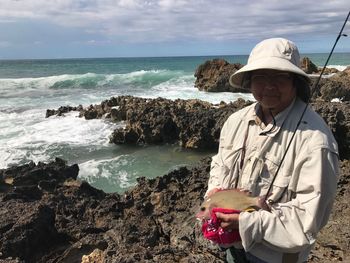 Portrait of senior woman holding fishing rod while standing at beach against sky