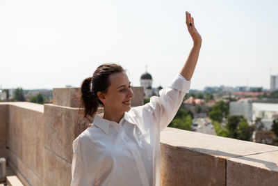 Young woman with arms raised standing against railing against clear sky