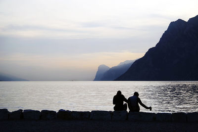 Silhouette couple sitting at lakeshore against sky during sunset