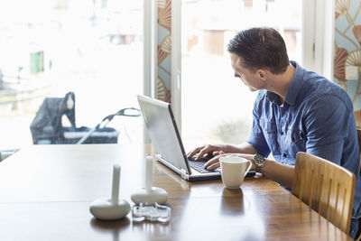 Father looking at baby carriage while using laptop at dining table