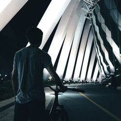 Rear view of man standing with bicycle on illuminated bridge at night