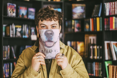 Portrait of young man with curly hair covering face with book in bookstore