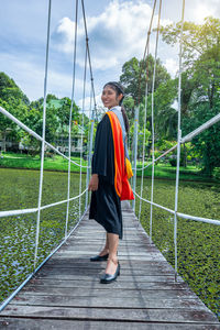Portrait of young woman wearing graduation gown standing on footbridge at park
