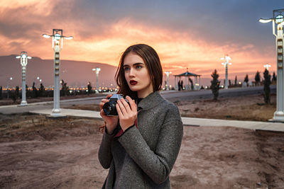 Portrait of young woman photographing against sky during sunset