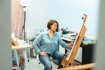 Concentrated middle age caucasian woman artist drawing with pencil on canvas at home art studio