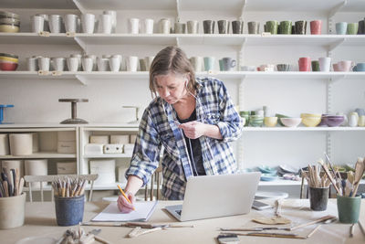 Mature female potter writing on paper while standing by laptop at table in workshop