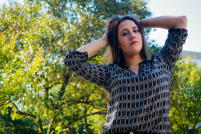 Portrait of woman with hands behind head standing against trees at park