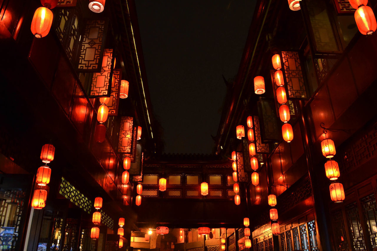 LOW ANGLE VIEW OF ILLUMINATED LANTERNS HANGING AMIDST BUILDINGS IN CITY