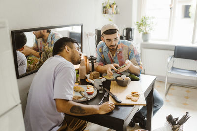 Gay couple sharing smart phone with each other while having food on dining table at home