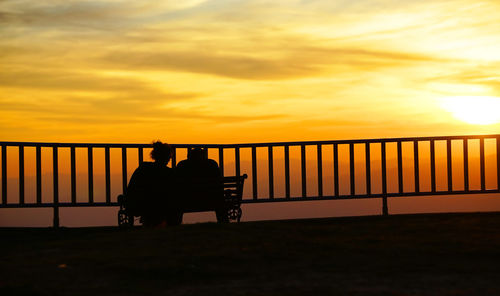 Silhouette people sitting on bench against orange sky
