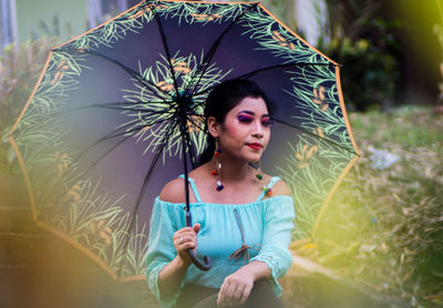 Young woman holding umbrella looking away sitting against plants