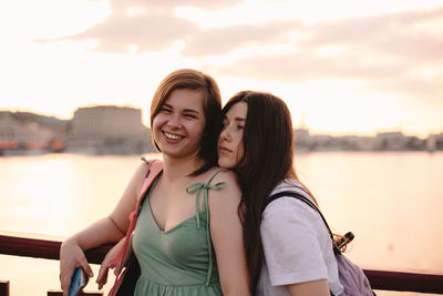 Two happy girlfriends standing on bridge against river at sunset