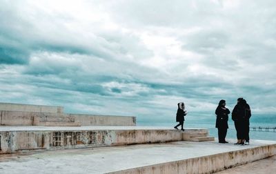 People standing on retaining wall by sea against sky