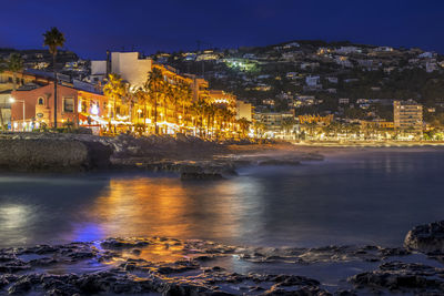 Illuminated buildings by sea against sky at night