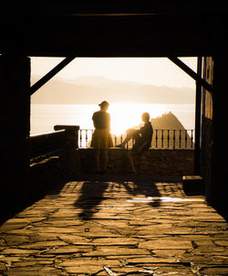 Men sitting on railing by sea against sky during sunset