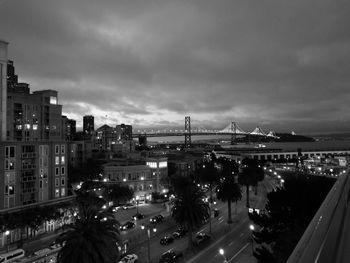 Illuminated city by bay against cloudy sky