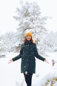 Cheerful girl with red hair in warm clothes playing with snow outdoors near the beautiful forest