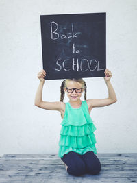 Smiling girl holding blackboard with text while kneeling against wall