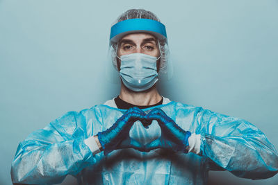 Portrait of doctor wearing mask making heart shape while standing against wall