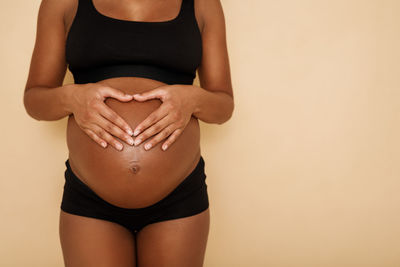 Midsection of pregnant woman with hands on stomach standing against beige background