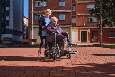 Caring nurse in uniform pushing elderly disabled woman in wheelchair while walking walking on paved sunny street near buildings in city