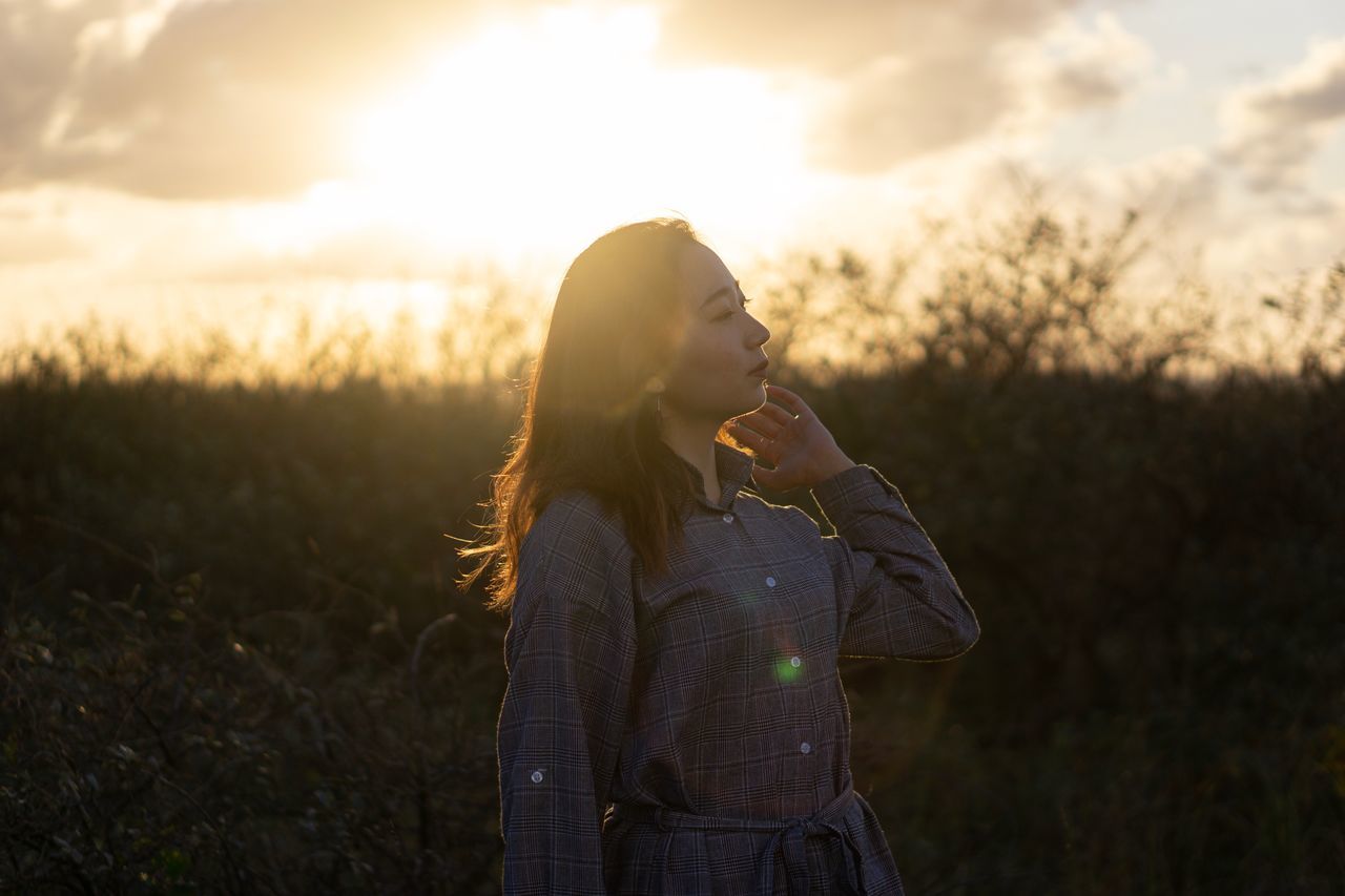 real people, one person, leisure activity, lifestyles, young adult, sky, young women, land, sunset, nature, standing, focus on foreground, casual clothing, waist up, field, looking, sunlight, adult, beautiful woman, lens flare, hairstyle, outdoors, contemplation, teenager