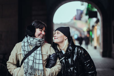 Smiling young female friends looking each other face to face while standing on street in city during winter