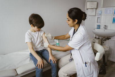 Female pediatrician disinfecting boy's arm sitting on bed at healthcare center