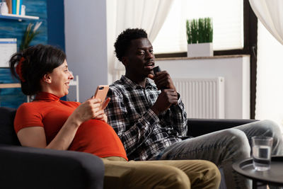 Man singing sitting by pregnant woman holding mobile phone on sofa