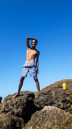 Low angle view of young shirtless man standing on rock formation against clear sky