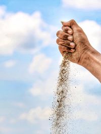 Cropped hand spilling sand against sky