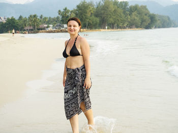 Portrait of woman wearing bikini standing at beach against trees