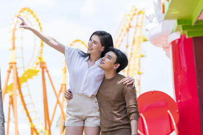 Couple in autumn outfit looking away near roller coaster in amusement park.