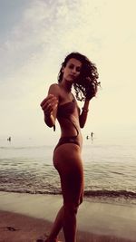Portrait of young woman standing at beach against sky during sunset
