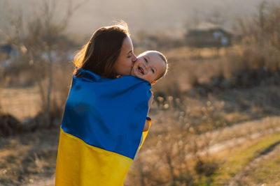 Woman hugs her little son wrapped in yellow and blue flag of ukraine in outdoors.