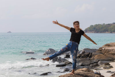 Full length portrait of woman with arms outstretched standing on rock at beach