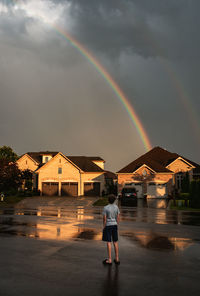 Boy standing in the street looking up at a rainbow in the sky.