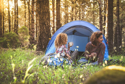 Smiling women holding coffee cups while sitting by tent in forest 