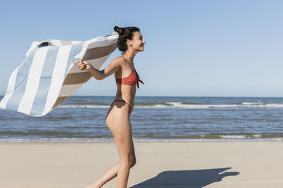 Young woman waving towel while running at beach during vacation