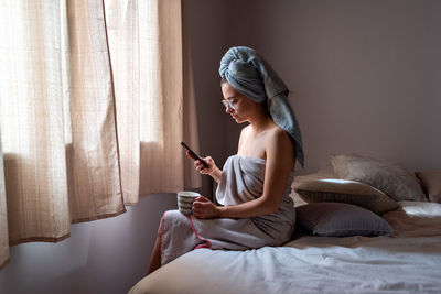 Young woman using mobile phone while sitting on bed at home