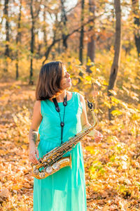 Girl with black hair standing in a yellow autumn park in a long blue dress with an alto saxophone