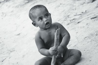 Naked baby boy holding wood while sitting on sand at beach