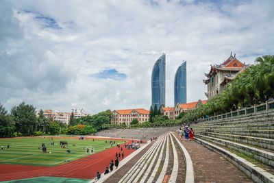 People at soccer stadium by modern buildings against cloudy sky