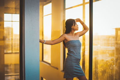 Woman wearing off shoulder dress standing by window at home during sunset