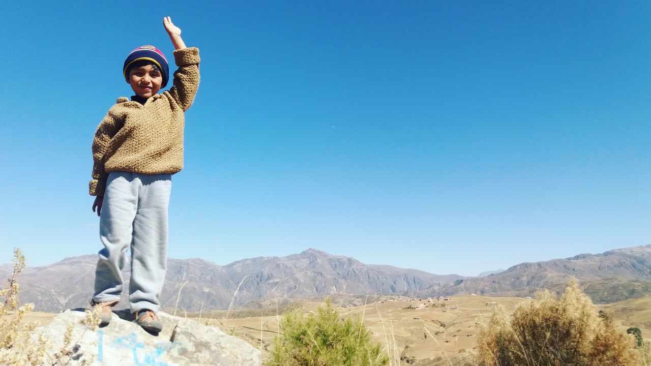 real people, clear sky, standing, mountain, one person, copy space, nature, day, outdoors, young adult, front view, lifestyles, young women, casual clothing, leisure activity, looking at camera, landscape, warm clothing, blue, happiness, full length, beauty in nature, mountain range, smiling, portrait, sky, people