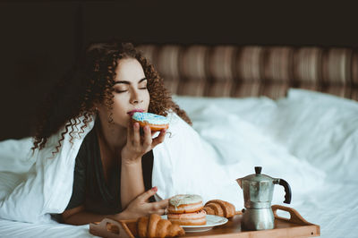 Woman having doughnut with eyes closed in bedroom