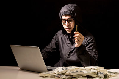 Computer hacker using laptop by paper currency at table against black background