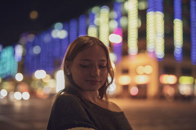 Portrait of smiling young woman standing against illuminated city at night
