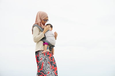 Mother wearing hijab carrying daughter against sky