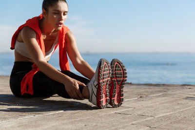 Young woman exercising while sitting on footpath against sea